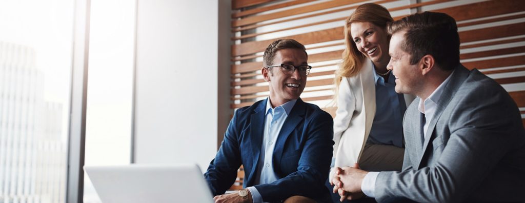 Dirección de Empresas Familiares. Dos hombres y una mujer, familiares y directivos de una empresa, están sentados frente a un laptop, riendo y discutiendo sobre los resultados positivos de la empresa.